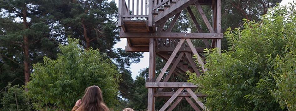 Twee kinderen wandelen over een zandpad richting een houten uitkijktoren, omringd door bomen en struiken.
