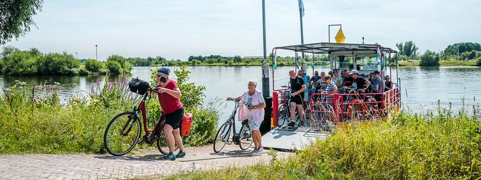 Radfahrer verlassen eine Fähre über die Maas auf einer der 5 Fahrradrouten und Tipps in Limburg, umgeben von Wasser und grüner Landschaft.