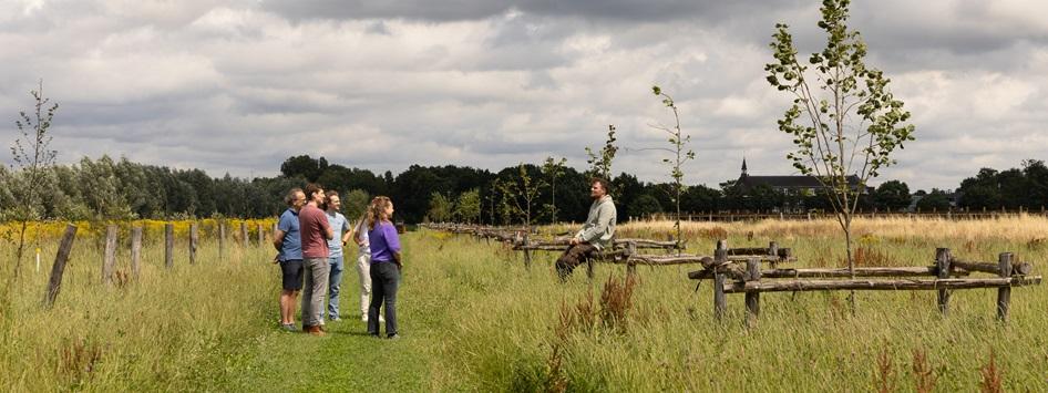 Een gids geeft een rondleiding aan een groep mensen in de groene omgeving van LEUKER in Heythuysen met weides en houten hekwerken, onder een bewolkte lucht. Perfect voor een dagje uit in Leudal