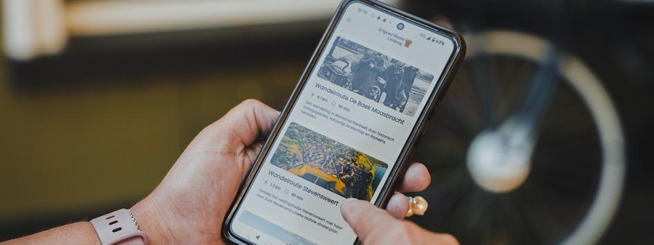 Persoon bekijkt wandelroutes op de Erfgoedroute Limburg app via een smartphone, met een fietswiel op de achtergrond.