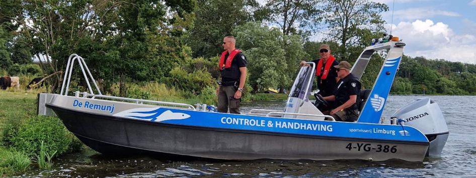 Een controle- en handhavingsboot van Sportvisserij Limburg, genaamd 'De Remunj', vaart langs de oever van een waterplas. Op de boot staan drie handhavers in zwarte uniformen en met reddingsvesten. Op de achtergrond grazen enkele runderen in een groen natuurgebied.