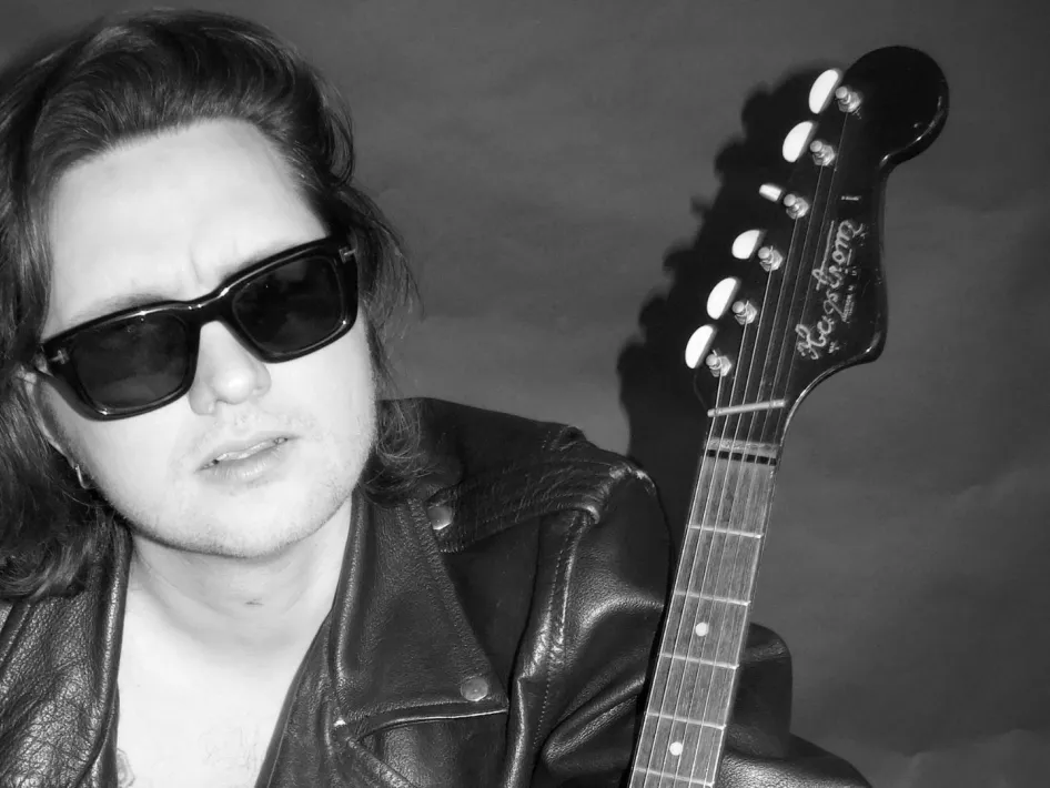 Jett Rebel met zonnebril en leren jas, poserend naast een elektrische gitaar in een zwart-witportret.