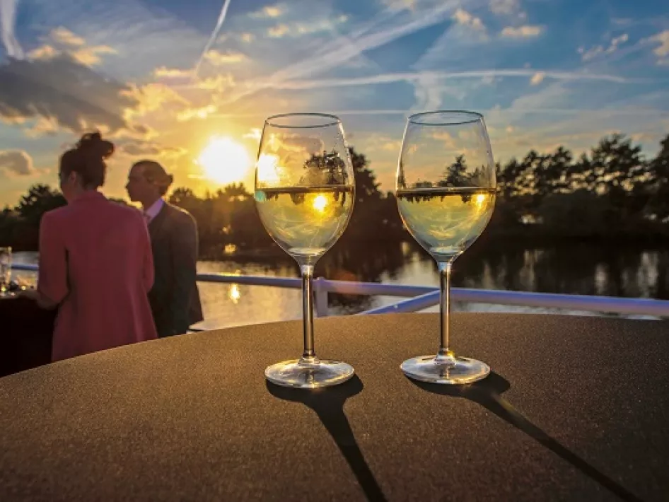 Twee glazen witte wijn op een tafel met een zonsondergang en gezellige gasten op de achtergrond.