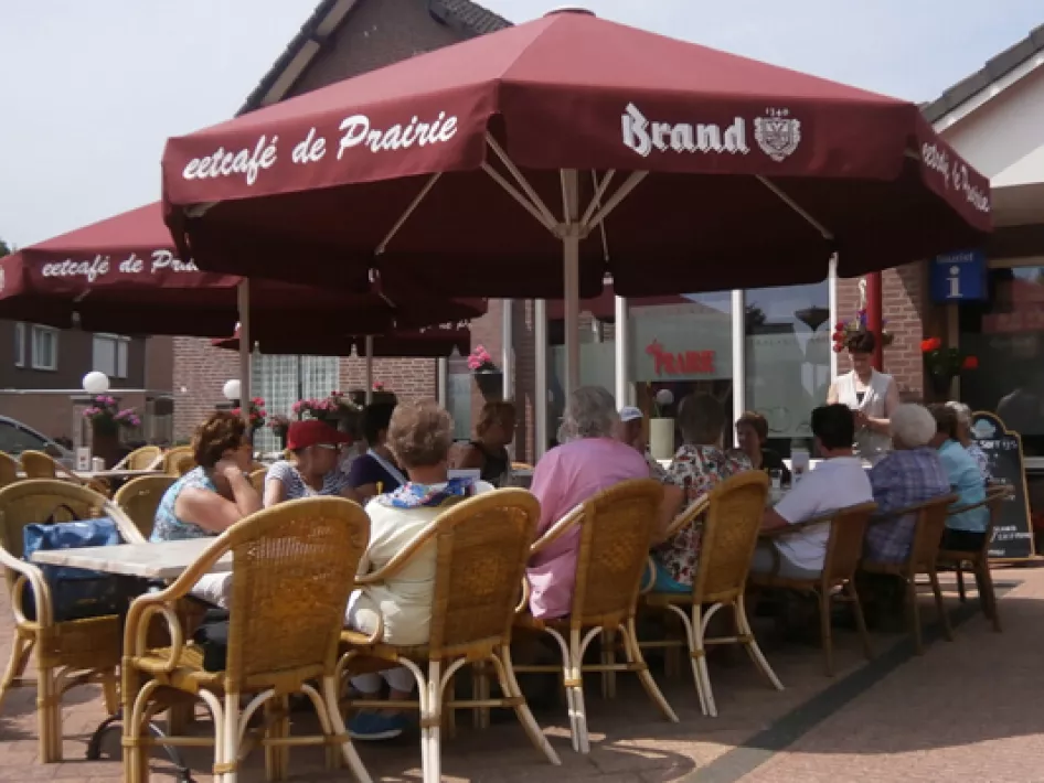 Gezellig terras bij Eetcafé De Prairie met gasten onder rode parasols, in gesprek en genietend van het buitenleven.