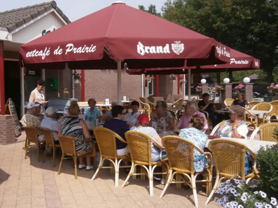 Groep mensen zit onder grote rode parasols op het terras van Eetcafé De Prairie, genietend van een drankje op een zonnige dag.