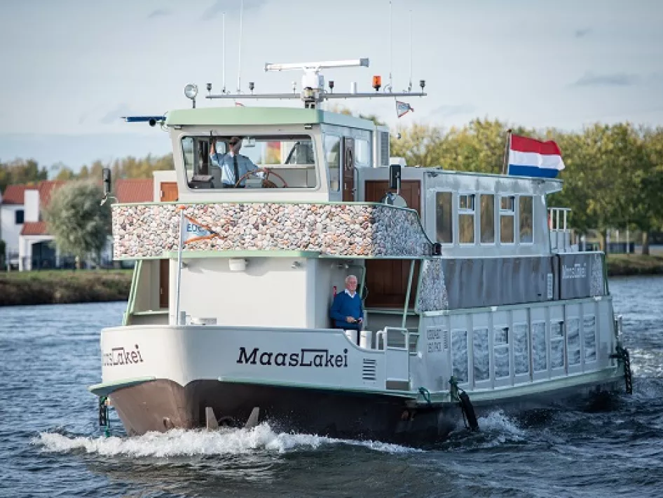 Passagiersschip MaasLakei vaart over het water met de Nederlandse vlag wapperend op de achtersteven.