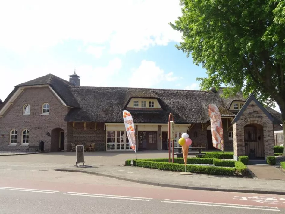 Buitenkant van een landelijke ijssalon met rieten dak, bakstenen gevel, vlaggen en een ijsco-display bij de ingang op een zonnige dag.