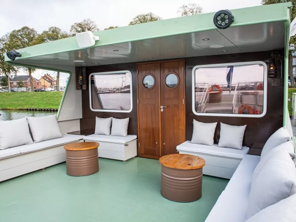 Overdekt buitendek van een schip met comfortabele loungebanken, houten tafels en nautische details.