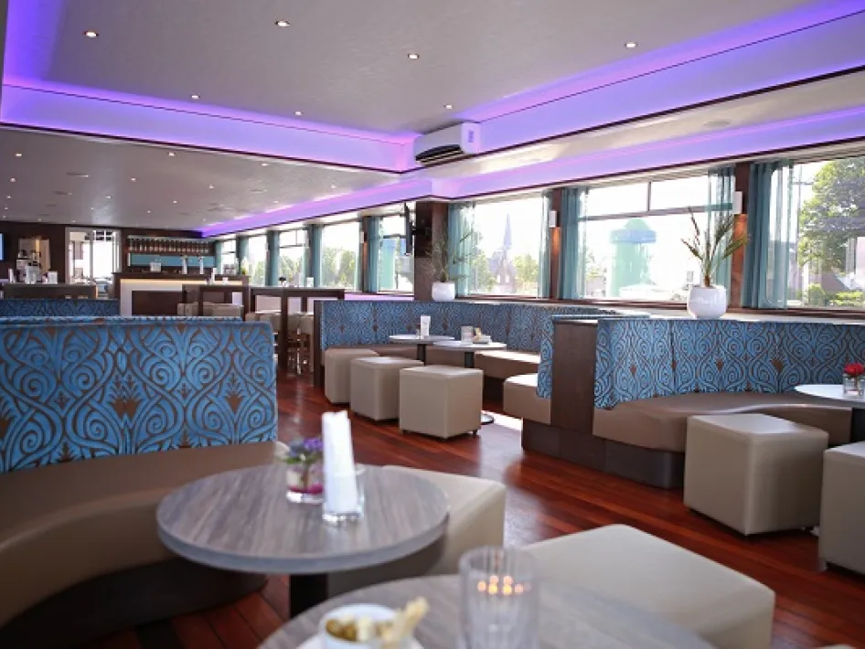 Sfeervolle lounge aan boord van een schip, met luxe zitbanken, houten tafels en paarse LED-verlichting.