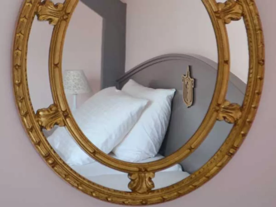 Ovale spiegel met gouden rand waarin een opgemaakt bed met wit linnengoed weerspiegeld wordt.