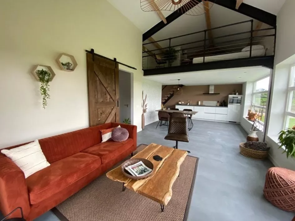 Sfeervol ingerichte woonkamer met een rode bank, houten salontafel, hoge plafonds en een vide met slaapgedeelte, uitkijkend op een open keuken.