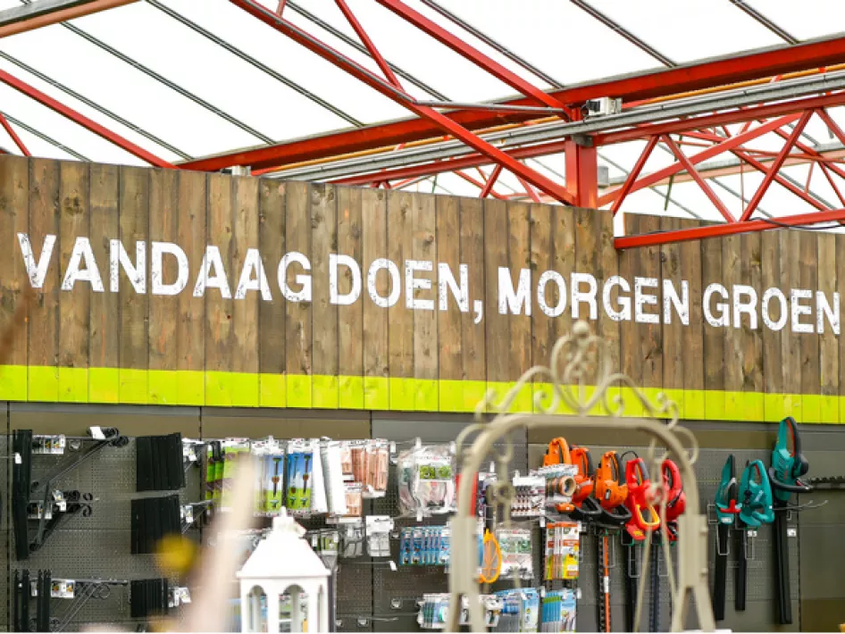 Een houten wand met de tekst "VANDAAG DOEN, MORGEN GROEN" in witte letters, onderdeel van een tuinwinkel met op de voorgrond diverse tuinartikelen en gereedschap, zoals snoeischaren en accessoires.