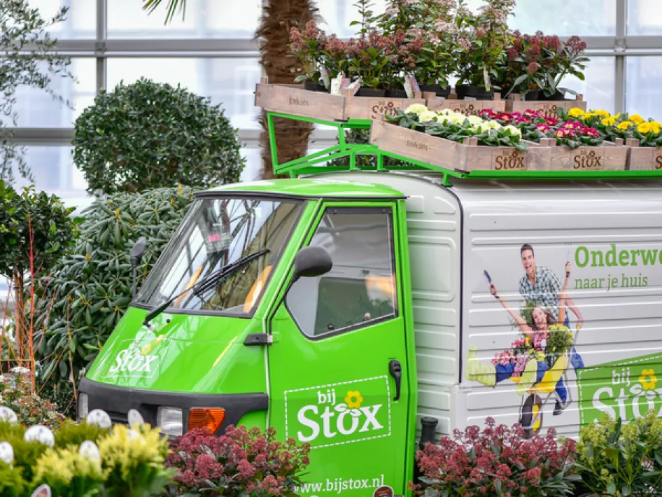 Een groene elektrische driewieler met het logo "bij Stox", omringd door kleurrijke bloemen en planten in een kas, met houten kisten vol bloemen op het dak en de slogan "Onderweg naar je huis" op de zijkant.