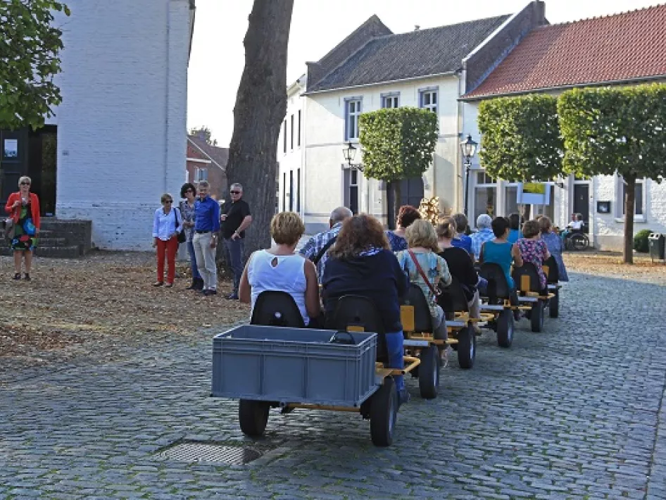 Group of people ride an electric train through a picturesque village with historic buildings.