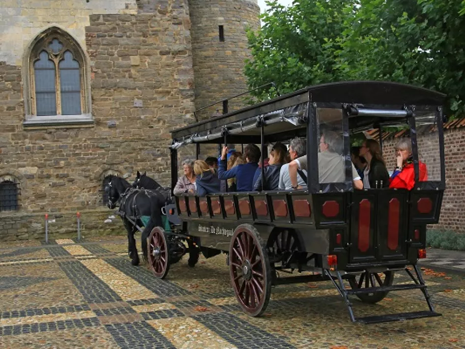 Horse-drawn streetcar with passengers rides along a cobblestone street past a historic building.