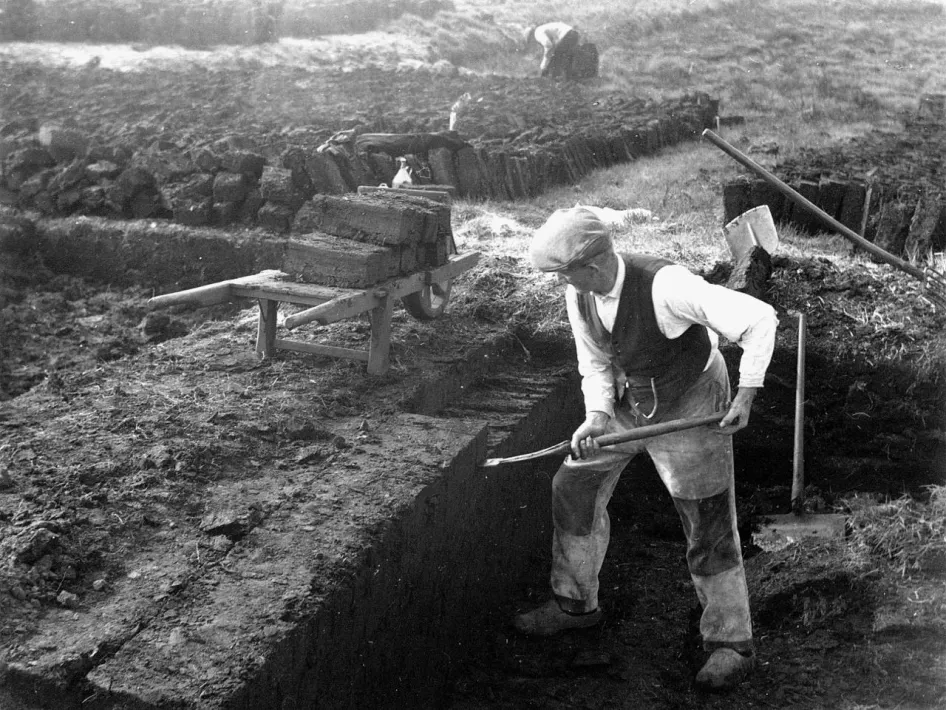 Historische foto van een turfsteker die turf afsteekt in De Groote Peel, met op de achtergrond turfvelden, gestapelde turven en een kruiwagen.