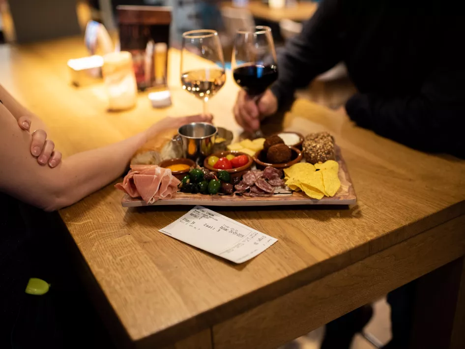 Tapas board is on the table while people toast with a glass of white and a glass of red wine.