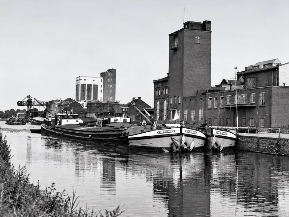 Historische zwart-witfoto van schepen op de Zuid-Willemsvaart bij industriële gebouwen in Weert, passend bij De vaart erin – 200 jaar Zuid-Willemsvaart.