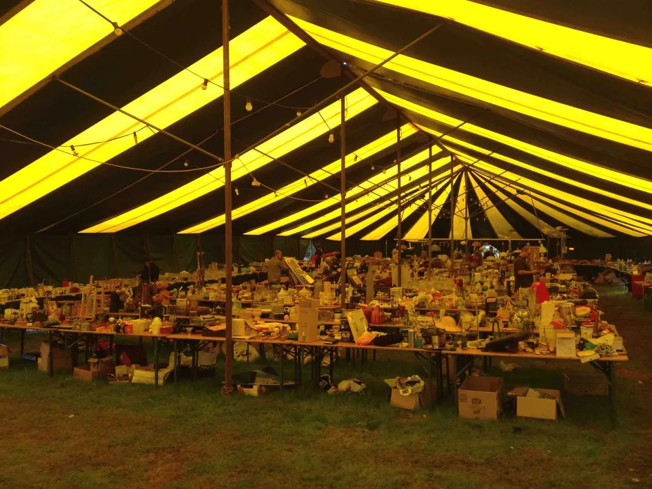 Tafels vol tweedehands spullen onder een grote geel-zwarte markttent op een vlooienmarkt.
