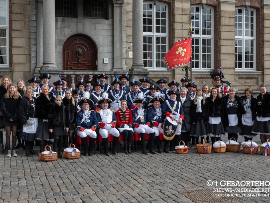 Groepsfoto van leden van een schutterij in traditionele uniformen, poserend voor een historisch gebouw. Voor hen staan manden op de grond, en naast de groep staan vrouwen in zwart-witte kleding met manden.