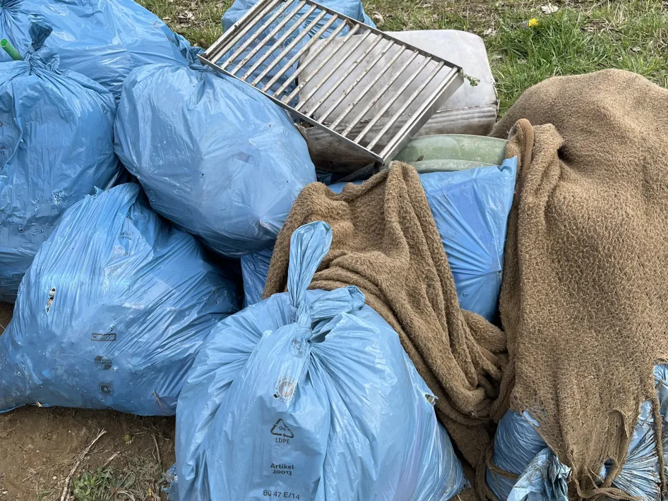 Blauwe vuilniszakken met afval liggen op een grasveld, met een metalen rek en een oude deken bovenop naast de Maas.