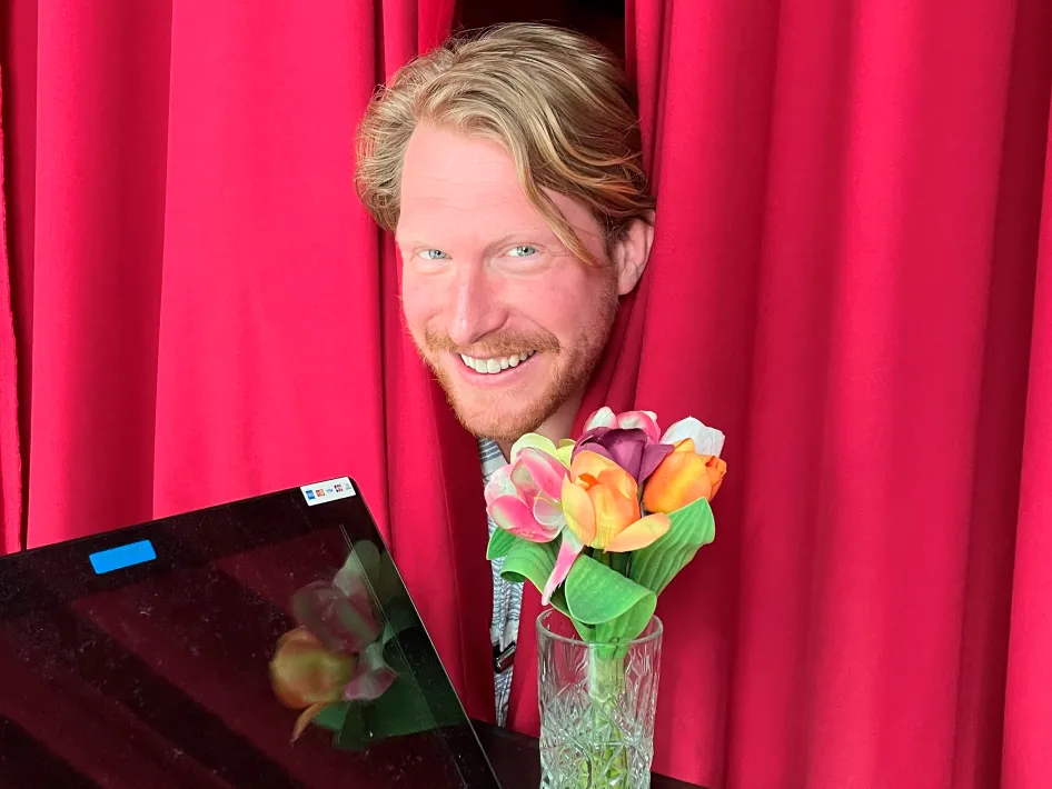 Kor Hoebe steekt zijn hoofd tussen rode theatergordijnen door met een klein bosje bloemen in een klein vaasje voor zich.