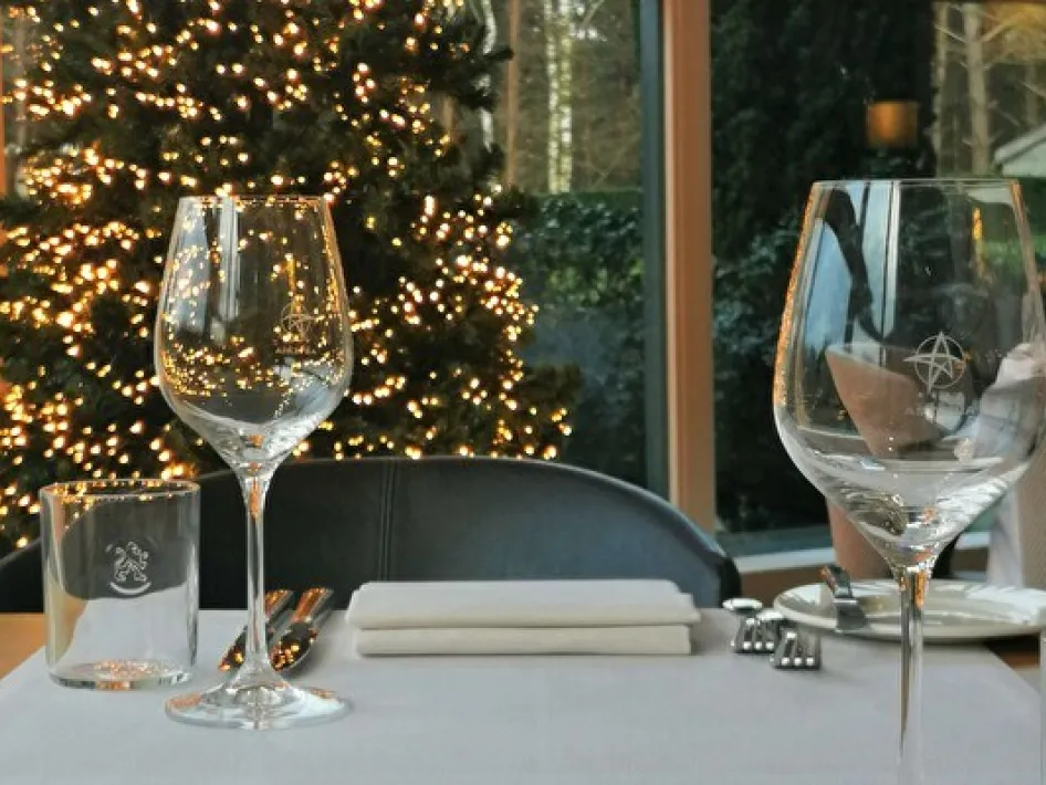 Gedekte restauranttafel met wijnglazen en servet, voor een raam met uitzicht op een verlichte kerstboom in sfeervolle omgeving.