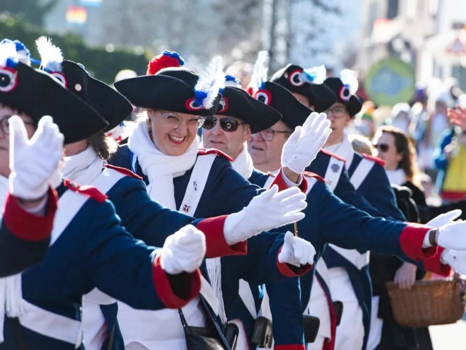 Leden van een schutterij in blauw-witte uniformen met zwarte hoeden en rode details, marcherend tijdens een optocht. Een vrouw in het midden lacht en zwaait enthousiast naar het publiek.