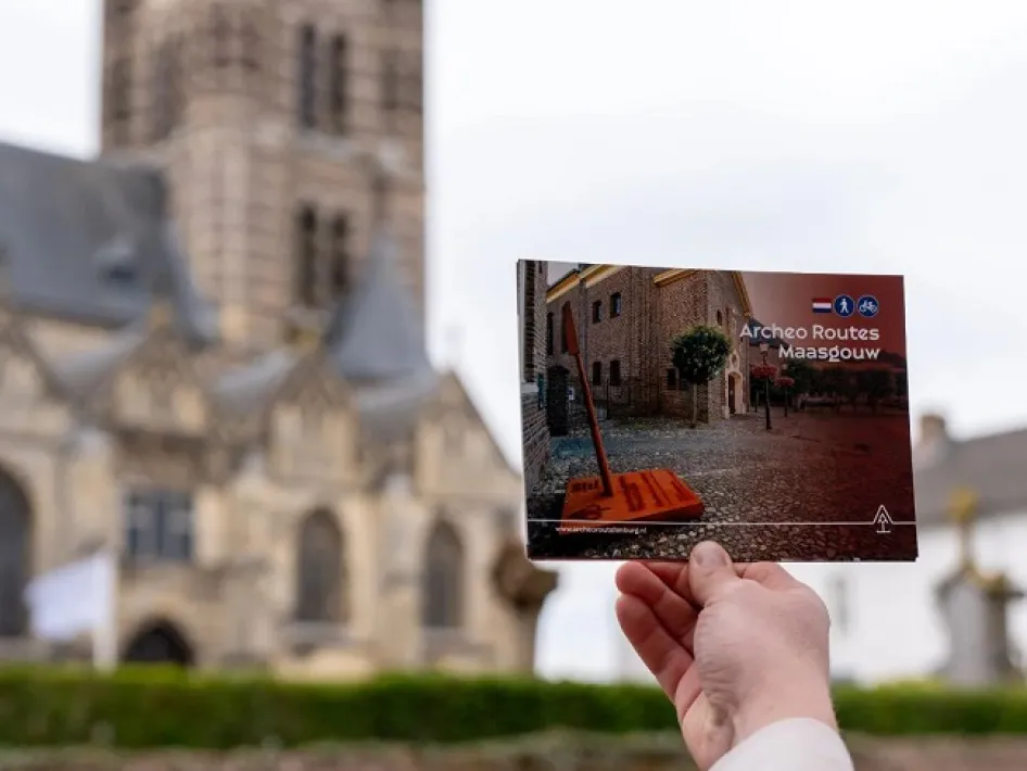 Persoon houdt brochure van Archeo Routes Maasgouw omhoog, met de kerk van Thorn op de achtergrond