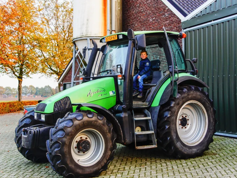 Jongen zit in een grote groene tractor op het erf van de boerderij.