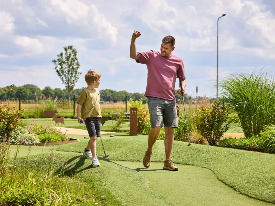 Vader viert een geslaagde slag met zijn zoon op de Adventure golfbaan van het park, omgeven door bloemen en gras.
