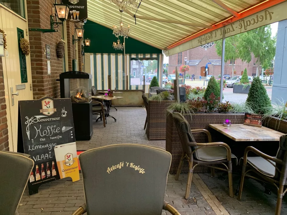 Covered and cozy terrace of an eatery, with comfortable rattan chairs, wooden tables and a fireplace for extra warmth. A chalkboard at the entrance offers specials such as coffee with Limburg flan. The village square is visible in the background.