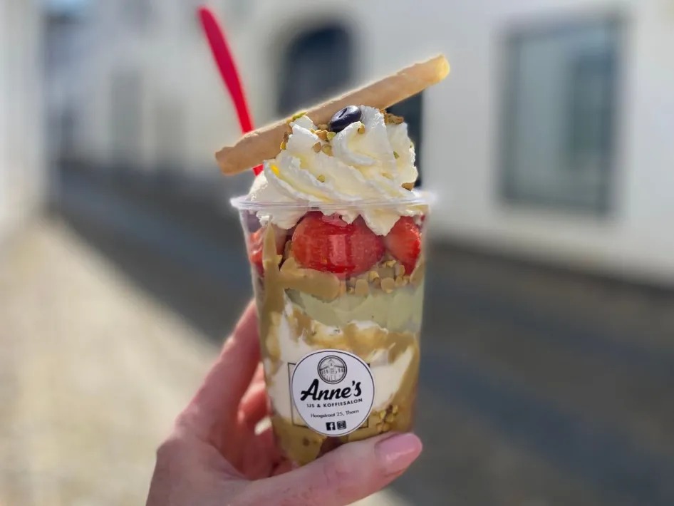 Close-up van een kleurrijke ijsbeker van Anne's IJs & Koffiesalon, gevuld met slagroom, aardbeien, nootjes en een wafelstaafje.