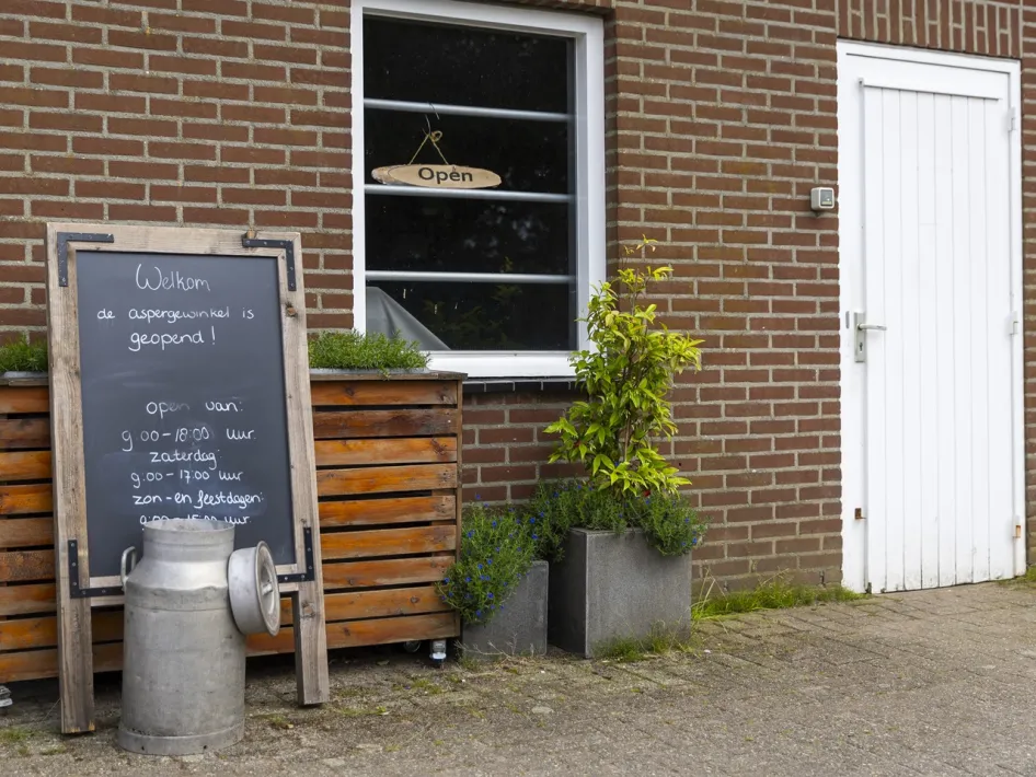 Boerderijwinkel Oppe Haes met een krijtbord waarop openingstijden staan, bij de ingang van een landelijke boerderij
