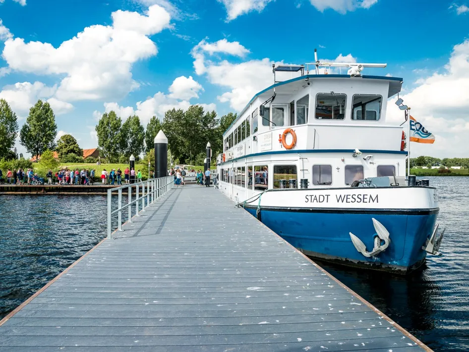 Rondvaartboot Stadt Wessem aangemeerd aan de steiger op de Maasplassen, waar bezoekers aan boord gaan voor de Asperge Cruise Voilà onder een blauwe lucht met wolken