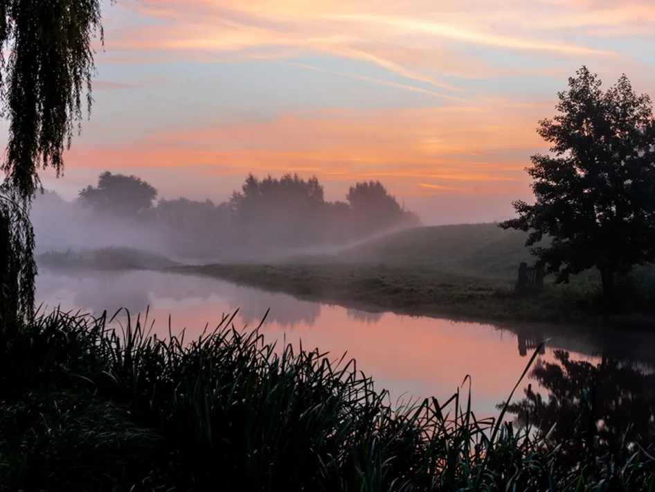 Blick auf einen ruhigen Fluss bei Sonnenaufgang, mit Nebel über dem Wasser und Silhouetten von Bäumen – in der Umgebung des B&B den Engel.