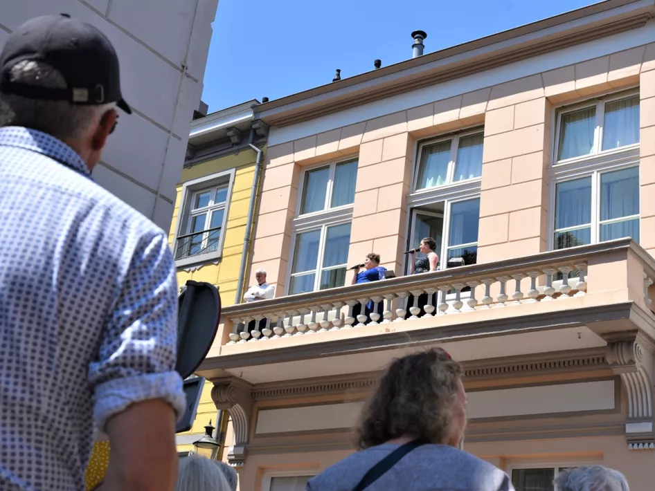 Mensen kijken vanaf een balkon naar een evenement op straat, terwijl voorbijgangers omhoog kijken naar het gebouw op een zonnige dag.