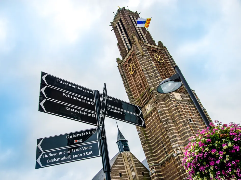 Blik omhoog naar de toren van de Sint Martinuskerk in Weert, met wegwijzers op de voorgrond en bloemen rechts in beeld.