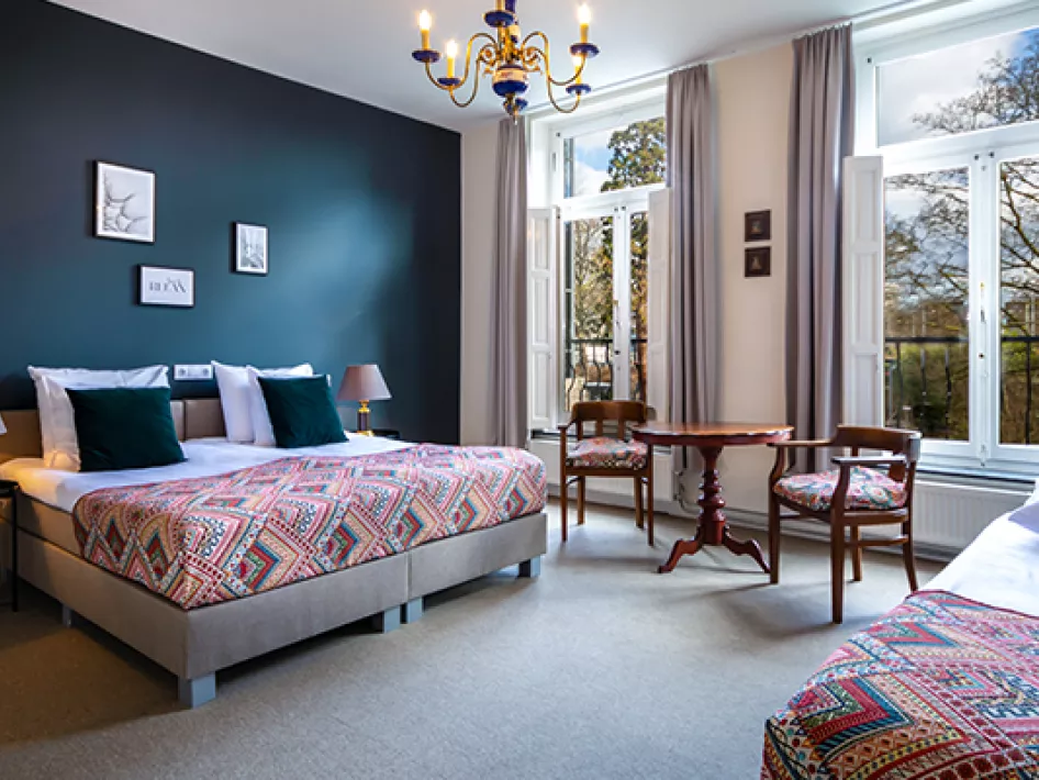 A stylish hotel room with a modern design, furnished with comfortable box-spring beds with colorful bedspreads, a small wooden table with chairs, and large windows overlooking green surroundings. The room has an attractive blue wall and is lit by a classic chandelier.