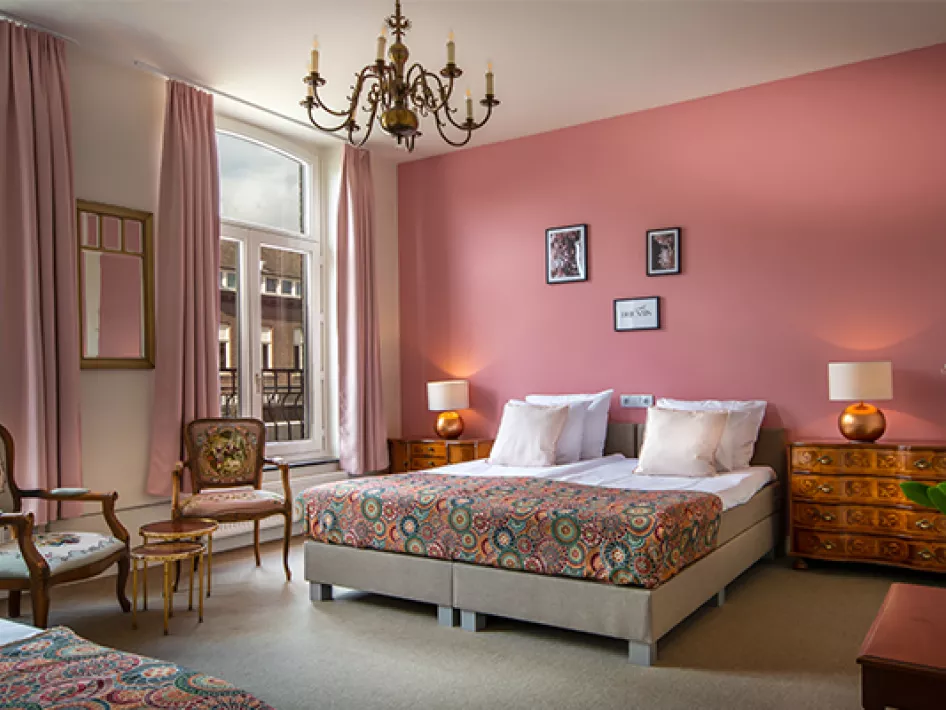 A stylish hotel room with a modern design, furnished with comfortable box-spring beds with colorful bedspreads, a small wooden table with chairs, and large windows overlooking a surrounding area. The room has an atmospheric pink wall and is lit by a classic chandelier.