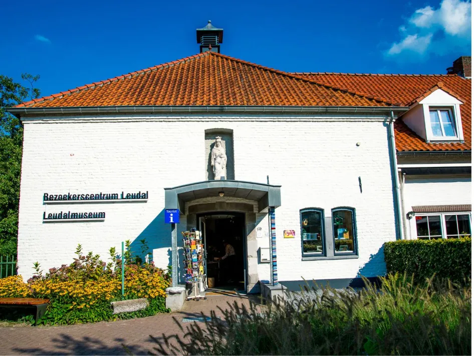 Facade of Visitor Center Leudal and Leudal Museum, a white building with red tiled roof, surrounded by flowers and greenery. At the entrance is an information board for tourists.
