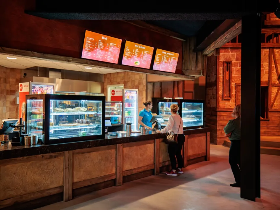 Besucher bestellen Essen und Getränke an einem Tresen mit beleuchteten Menütafeln in einem Indoor-Gastronomiebereich.