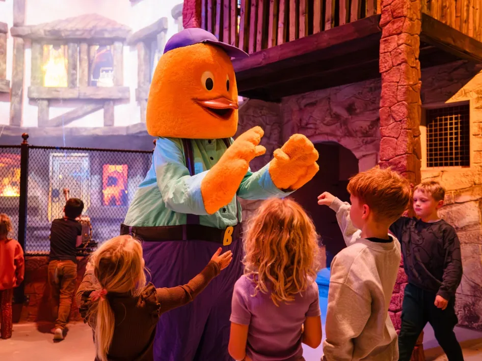 De mascotte Billy in een blauw shirt en paarse pet begroet kinderen in het indoor themagebied.