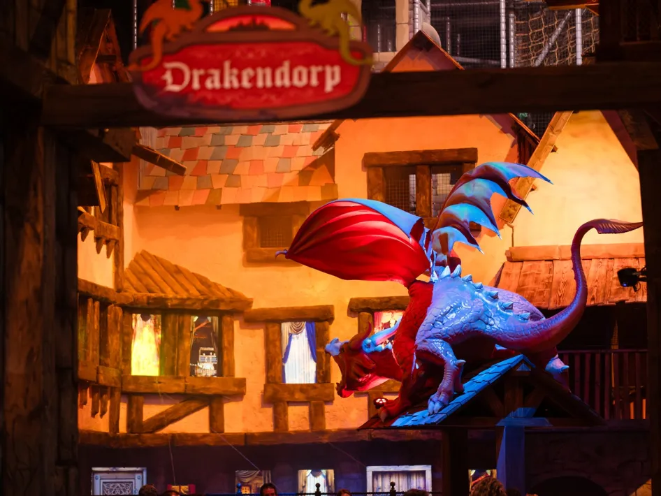 Een grote draak staat op een dak in Drakendorp bij BillyBird Drakenrijk met een brandend dorp op de achtergrond.