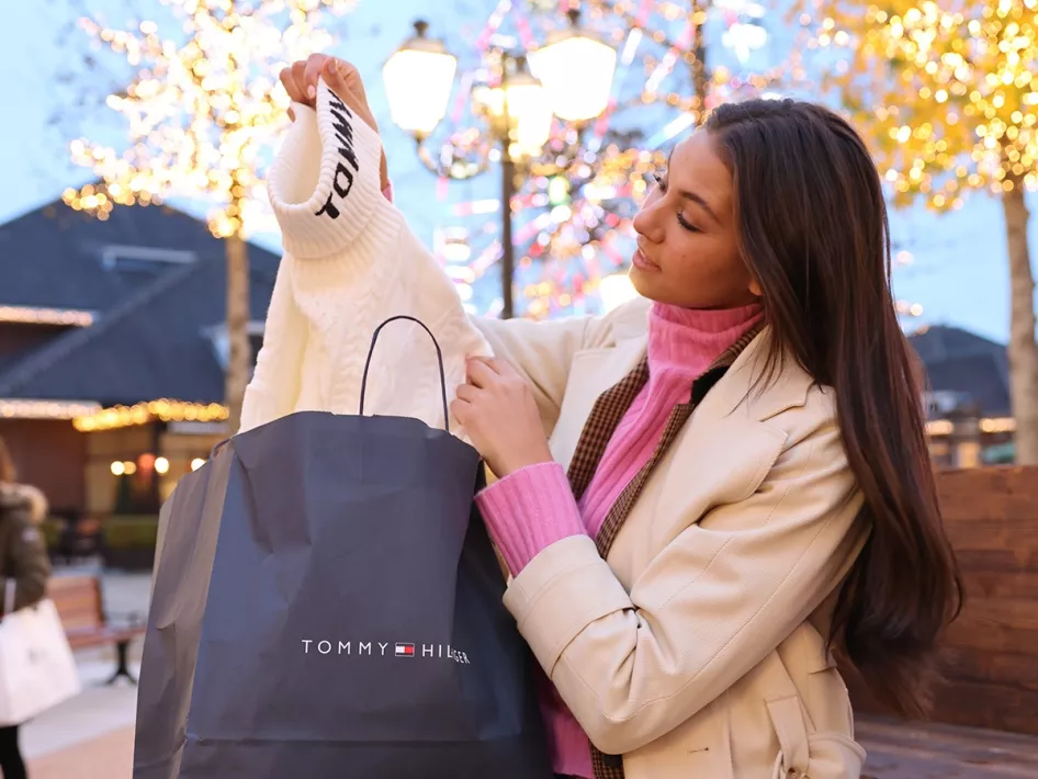 Woman takes a white sweater from a Tommy Hilfiger shopping bag during the Black Friday Days at Designer Outlet Roermond, surrounded by festive holiday lights.