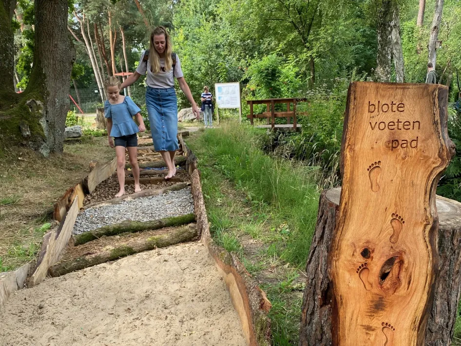 Volwassene en kind lopen op blote voeten over een blotevoetenpad met zand, grind en dennenappels bij het Natuur- en Milieucentrum in Weert, met houten bord waarop ‘blote voeten pad’ staat.