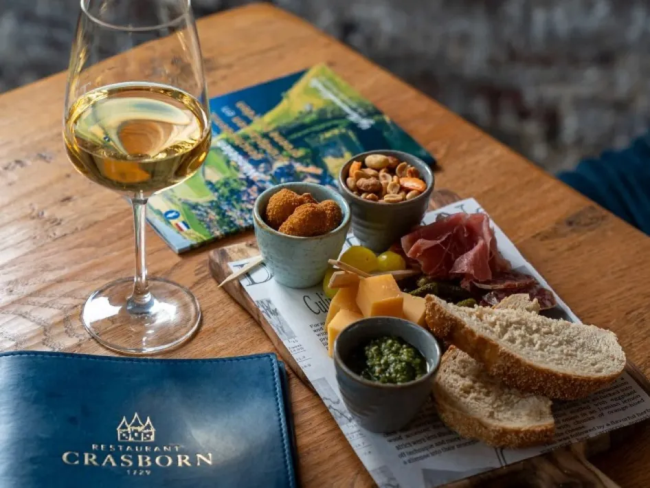 Borrelplank met brood, kaas, ham en witte wijn op tafel, naast het menu van Crasborn