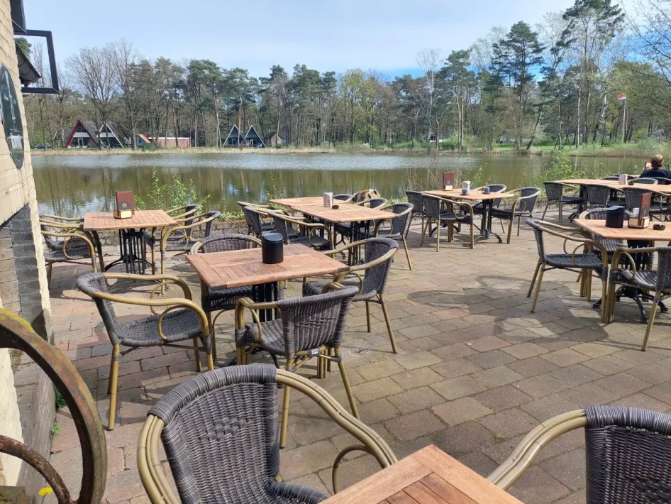 Open terras met rieten stoelen en houten tafels, direct aan het water gelegen met zicht op het bosrijke vakantiepark.