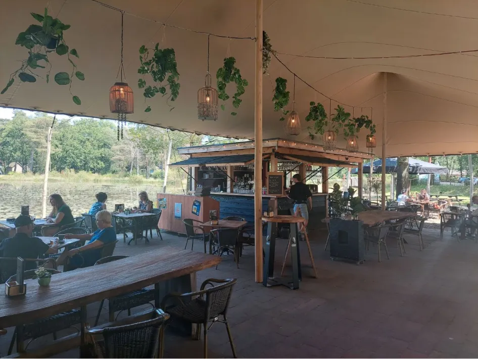 Gemoedelijk terras met houten tafels, hangplanten en uitzicht op het water, waar gasten ontspannen zitten