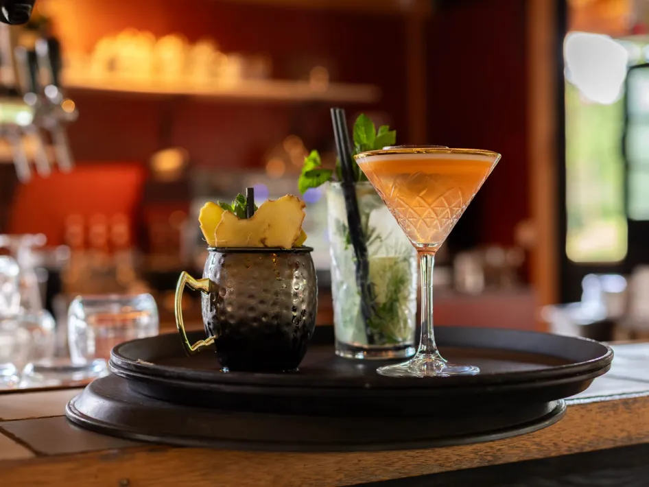 Tablett mit drei Cocktails an der Bar bei Boshuys Nieuw Vosseven, mit einem Kupferbecher mit Ingwer, einem mojitoähnlichen Getränk mit Minze und einem Cocktailglas vor warmem Restauranthintergrund